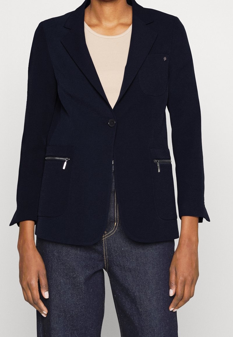 Blazer bleu marine ajusté en tissu léger, doté d'un bouton unique, de deux poches frontales zippées et d'un col cranté.