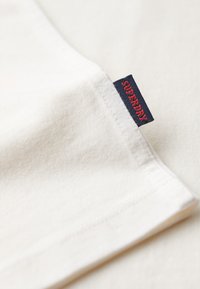 Tissu blanc avec une étiquette bleu marine cousue dans la couture, portant le texte rouge "SUPERDRY" en lettres majuscules.