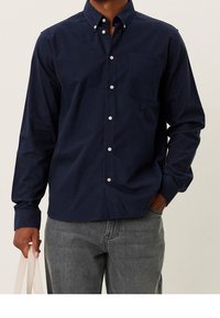 Camicia blu navy a maniche lunghe con bottoni, in cotone, con colletto, tasca frontale e bottoni bianchi, abbinata a jeans grigi e una borsa tote in tela.