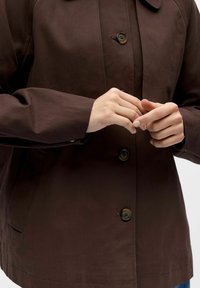 Personne portant une veste marron foncé boutonnée, en train d'attacher ou d'ajuster les boutons près de la poitrine avec les deux mains.