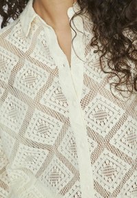 Chemise blanche légère à boutons, présentant un motif complexe en forme de losange. Le tissu est transparent et texturé, avec des détails subtils autour du col.