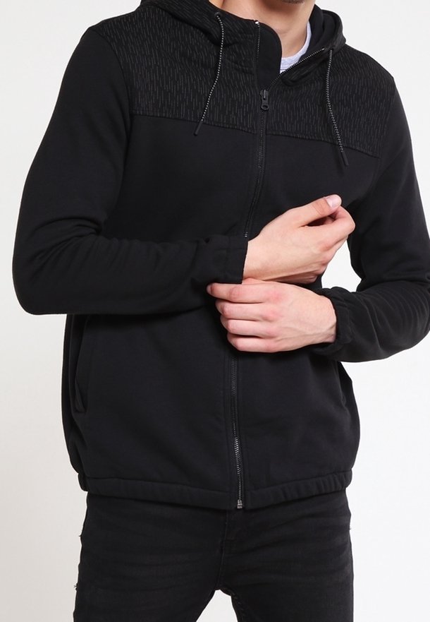 Homme portant un sweat à capuche noir zippé avec un motif sur le haut de la poitrine et la capuche, ajustant la manche gauche de la main droite.