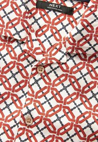 Kortærmet skjorte med rød geometrisk mønster på hvidt stof, beige knapper og mærkeetiket "NEXT Made in Bangladesh."