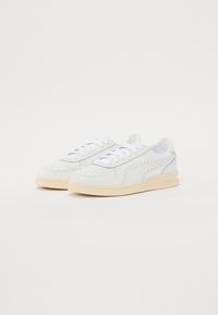Zapatillas blancas hechas de cuero y material sintético, con detalles perforados, cordones planos blancos y una suela de goma crema.