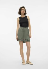 Top negro sin mangas combinado con pantalones cortos verde oliva que presentan bordados florales en el dobladillo. La modelo lleva zapatos de color claro con tiras en el tobillo.