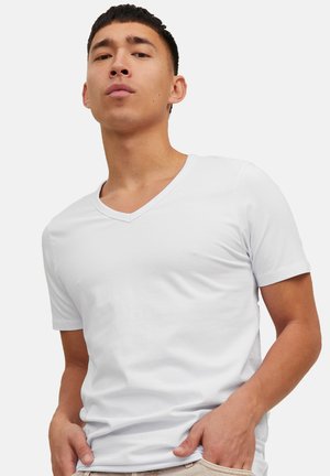 Jonge man die een effen witte T-shirt met V-hals en korte mouwen draagt, staand met handen bij de zakken tegen een effen achtergrond.