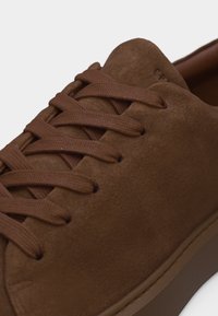 Καφέ σνίκερ από σουέντ με καφέ επίπεδα κορδόνια, κοντινή λήψη που δείχνει την υφή της επιφάνειας και την άκρη της λαστιχένιας σόλας.