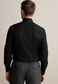 Schwarzes Langarmhemd mit spitzem Kragen, mit glatter Textur und sichtbaren Knopfdetails an den Manschetten, getragen mit grauen Hosen.