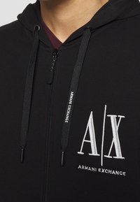 Sweat à capuche zippé noir avec cordons, avec texte blanc "Armani Exchange" et broderie du logo "A|X" sur la poitrine.