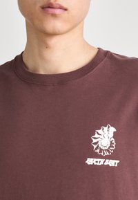 Hnědé bavlněné tričko s bílou grafiku mušle a textem "EARTH BEAT". Kulatý výstřih, krátké rukávy, hladká textura.