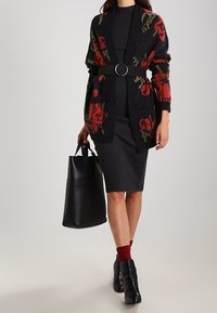 Cardigan negro con estampado floral, cinturón y vestido negro acanalado. Bolso de cuero negro, calcetas rojas y botas de tobillo.