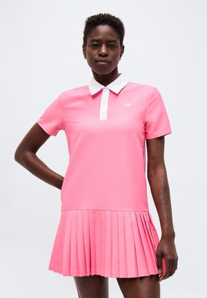 Mujer con un vestido polo rosa de manga corta, con cuello blanco y falda plisada, de pie con una mano en la cadera.