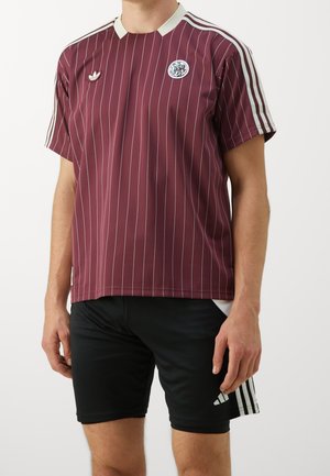 Mann trägt ein kastanienfarben gestreiftes Sporttrikot mit weißen Streifen auf den Schultern und schwarze Sportshorts mit weißen Streifen an den Seiten.