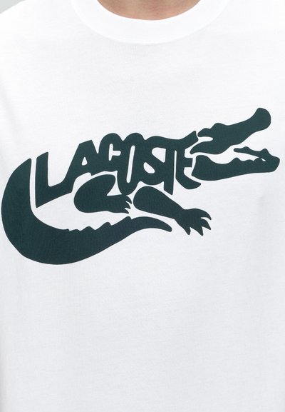 Черно стилизирано лого на крокодил с интегриран текст „Lacoste“, отпечатано на бяла предна част на риза.