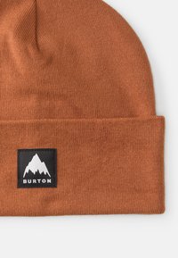 Brūns adīts cepure ar salocītu maliņu un melnu kvadrātveida uzlīmi, uz kuras attēlota balta kalnu dizaina un teksts "BURTON". Mīksta tekstūra.