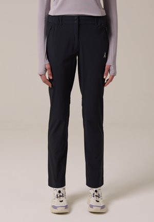 Persoon die zwarte rechte broek met zijzakken en witte sneakers draagt, staand tegen een effen achtergrond.