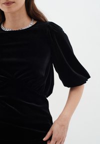Top in velluto nero con maniche a sbuffo, arricciato in vita, caratterizzato da un colletto geometrico argento. Tessuto liscio e design strutturato.