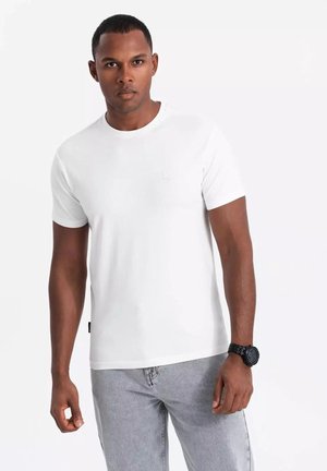 Vit kortärmad t-shirt med rund halsringning, tillverkad av bomull. Har en liten logotyp på bröstet och texturerade grå jeans med en mörk klocka.