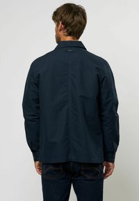 Chaqueta ligera de color azul marino con collar clásico, mangas largas y diseño de espalda recta. Presenta puños con botones y un logo sutil.
