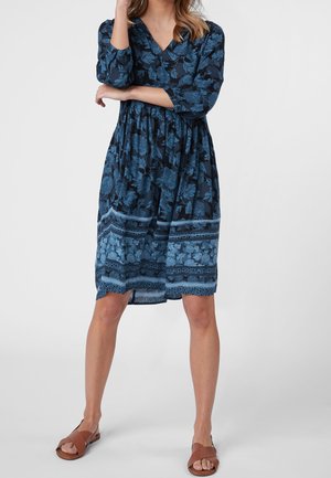 Femme portant une robe bleue à fleurs jusqu'aux genoux avec des manches trois-quarts et des sandales marron à bout ouvert, debout les bras croisés.