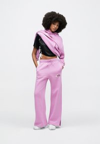 Ροζ φόρμες με κορδόνι στη μέση, μια αντίστοιχη φαρδιά μπλούζα με κουκούλα που είναι περασμένη στους ώμους, και ένα μαύρο cropped top με υφή από δίχτυ.