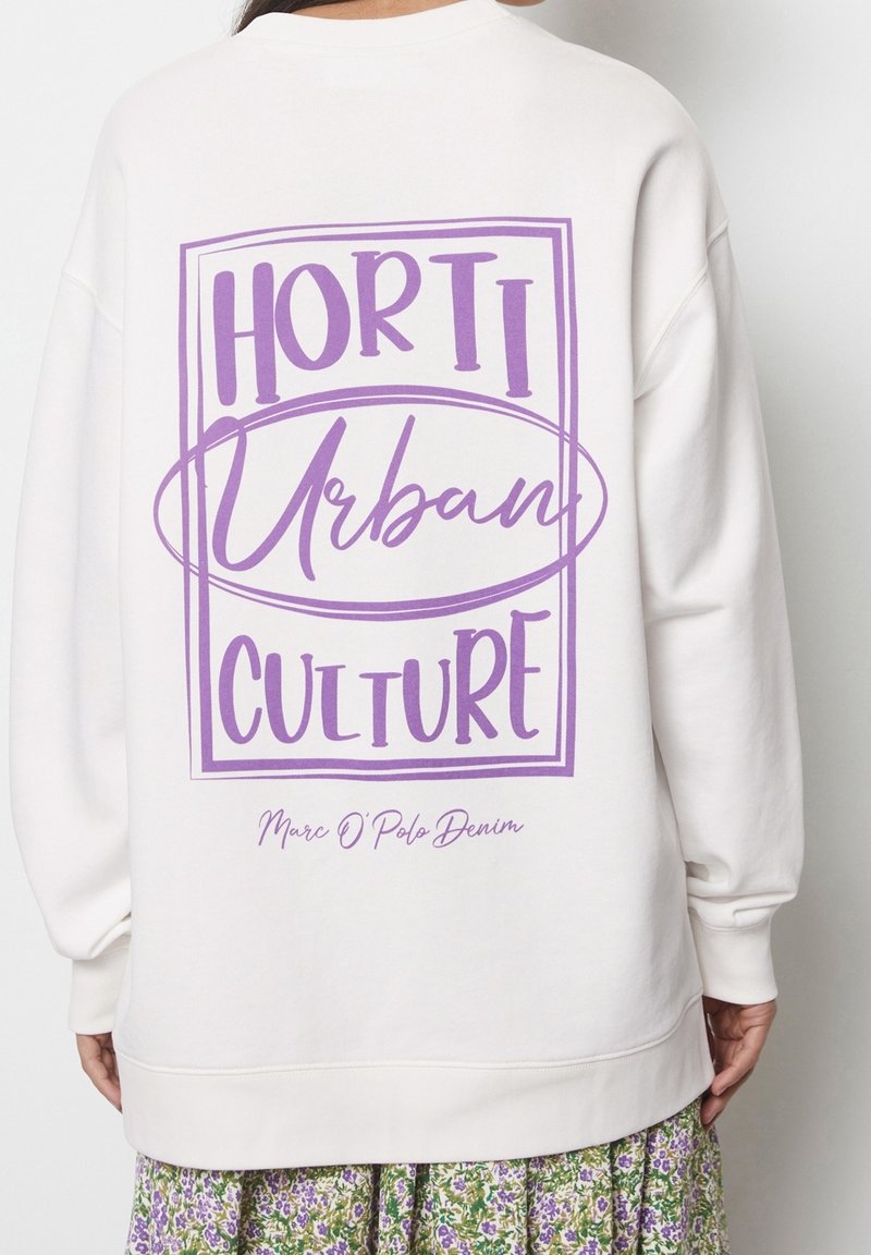 Sweat-shirt blanc avec texte violet "HORTI Urban CULTURE" encadré dans le dos, porté par une personne en jupe fleurie avec des fleurs vertes et violettes.