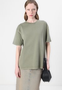 Olive green oversized t-shirt with short sleeves, crew neck, and casual fit, paired with a beige denim skirt, holding a small black handbag.