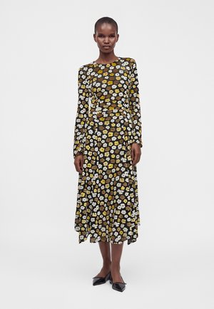 Femme portant une robe midi noire à manches longues avec un imprimé floral jaune, marron et blanc, et des ballerines noires pointues, debout devant un fond blanc.
