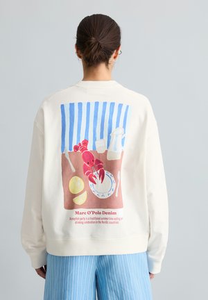 Femme portant un sweat-shirt blanc avec une illustration colorée d’écrevisses et de table dressée au dos, assorti à un pantalon bleu rayé.