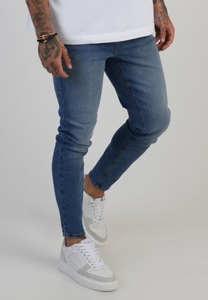 Homme portant un jean slim bleu, des baskets blanches et une chemise blanche avec des mains tatouées visibles sur un fond uni.
