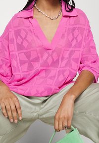 Rosa, texturerad stickad topp med V-hals och överdimensionerade ärmar, med ett geometriskt mönster. Modellen bär ljusgröna byxor och en matchande v äska.