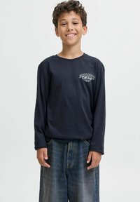 Leende pojke med kort lockigt hår iklädd en marinblå långärmad tröja med logotyp och löst sittande blekblå jeans mot en enkel bakgrund.
