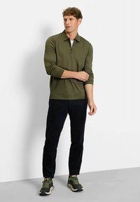 Olive green long-sleeve polo shirt with a half-zip collar, paired with black ribbed trousers and dark athletic shoes with a textured sole.