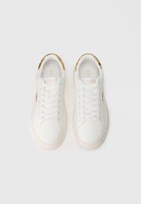 Ein Paar weiße Schnürsneaker mit goldenen Akzenten an der Ferse und an der Seite, mit dem Markennamen "GUESS," von oben auf weißem Hintergrund dargestellt.