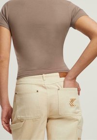 Osoba ubrana w taupe dopasowany t-shirt z krótkim rękawem oraz kremowe szerokie spodnie z tylnymi kieszeniami, jedna ręka w kieszeni, jasne neutralne tło.