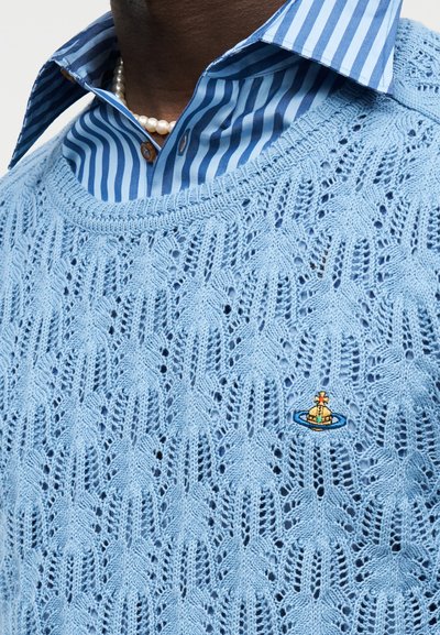 Pull en maille torsadée bleu clair porté sur une chemise à col rayée bleue, avec un collier de perles et un petit emblème brodé sur le pull.