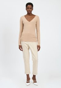 Beige V-Ausschnitt-Pullover mit langen Ärmeln, kombiniert mit hellen, umgeschlagenen Hosen und weißen, spitz zulaufenden Schuhen mit einem dekorativen Riemen.