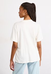 T-shirt en coton de couleur crème avec des manches courtes et un col rond ; la vue arrière révèle un design simple et une coupe décontractée. Pantalon bleu clair présenté.