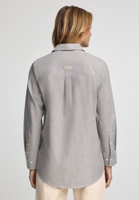 Ljusgrå knäppskjorta gjord av mjukt tyg, med ryggsömmar, rundad nederkant och dubbelknäppning vid ärmarna. Subtil logotag på baksidan.