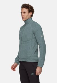 Giacca in pile di un verde bluastro opaco, con zip completa e colletto alto, due tasche frontali e una targhetta con logo sulla manica sinistra. Texture liscia.