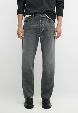 Mand iført løstsiddende, falmede sorte jeans, sorte sko og en sort langærmet top, stående mod en ensfarvet baggrund.