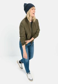 Olijfgroen bomberjack, donkerblauwe gebreide beanie, blauwe spijkerbroek, witte sneakers met grijze accenten, staand in een speelse pose.