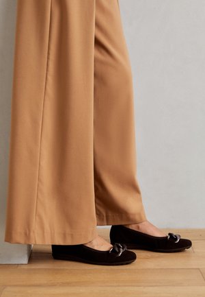 Czarne zamszowe baleriny z dekoracyjnym metalowym łącznikiem na palcach, w zestawieniu z szerokimi beżowymi spodniami na drewnianej podłodze.