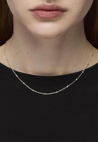 Collar de cadena de plata con pequeñas cuentas redondas, que presenta un diseño delicado y una textura suave, llevado sobre una camisa negra.