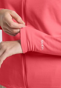 Rotes Sportshirt aus strukturierter Stoff, mit Frontreißverschluss und enganliegenden Ärmeln. Logo "TEXADRI" am Bündchen, das die Leistungsmerkmale des Designs präsentiert.