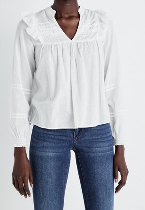 Donkere persoon gekleed in een witte blouse met lange mouwen en ruches, en blauwe spijkerbroek, staand tegen een effen lichte achtergrond.