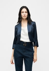 Femme portant un blazer court bleu foncé à manches trois-quarts, un haut blanc orné de dentelle et un pantalon taille haute bleu foncé avec une ceinture.