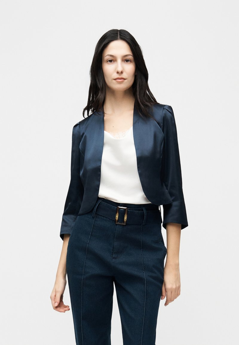 Femme portant un blazer court bleu foncé à manches trois-quarts, un haut blanc orné de dentelle et un pantalon taille haute bleu foncé avec une ceinture.
