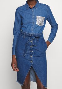 Denim shirt dress with a collar, long sleeves, button front, belted waist, and a silver sequined pocket. Features a high slit and hem accents.