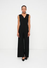 Mono negro con cuello drapeado, diseño sin mangas, piernas amplias y tela suave. Llevado con tacones negros de punta.